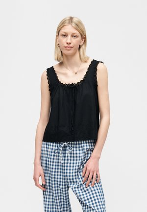 Noor naine, kes kannab varrukateta musta pitsiga kaunistatud topi ja sinise-valge ruudulise paelaga pükse, seisab ühevärvilise tausta ees.