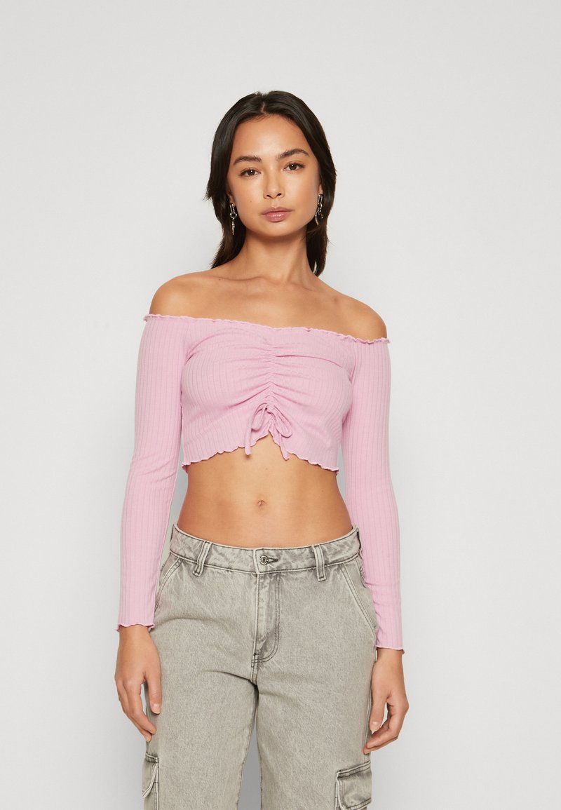 Ružičasti rebrasti crop top s dugim rukavima, off-shoulder dizajnom, nabranim detaljima i uvijenim čvorom sprijeda; valoviti rub dodaje teksturu.