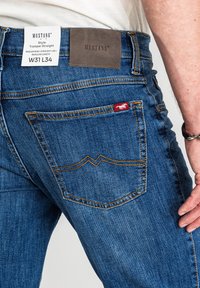 Niebieskie jeansy z denimu o średnim stanie i prostych nogawkach. Charakteryzują się kontrastowymi szwami, brązową skórzaną naszywką oraz małą czerwoną etykietą z logo.