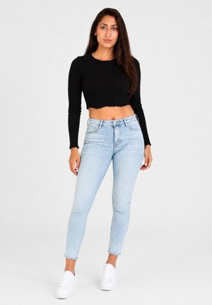 Jonge vrouw met lang bruin haar die een zwart crop top met lange mouwen draagt, lichtblauwe high-waisted jeans en witte sneakers, staand.