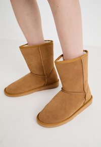 Sandfärgade mockaankelstövlar med en mjuk, krämfärgad foder. Designen har en platt sula och rund tå, med synliga sömnadsdetaljer.