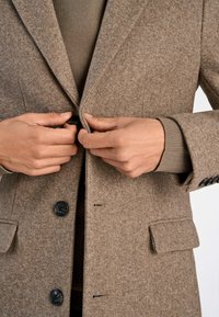 Manteau en laine brun mélangé avec un col cranté, des boutons noirs et des poches plaquées, présentant une finition texturée et douce. Des mains ajustent les boutons.