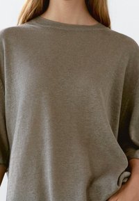 Oliivinvihreä, lyhythihainen neulepusero, jossa on pyöreä pääntie ja pehmeä, hieman teksturoitu kangas. Rentoa istuvuutta ja väljää siluettia.