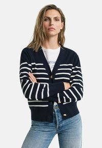 Marineblauer Cardigan mit weißen horizontalen Streifen, Knopfverschluss vorne und gerippten Bündchen. Kombiniert mit hellblauen, lässig geschnittenen Jeans.