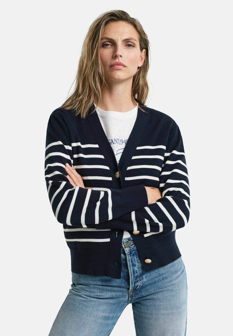 Marineblauer Cardigan mit weißen horizontalen Streifen, Knopfverschluss vorne und gerippten Bündchen. Kombiniert mit hellblauen, lässig geschnittenen Jeans.