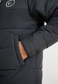 Black puffer jacket with quilted design and a visible Nike logo. Features side pockets and "THERMA-FIT" embroidery on the sleeve.