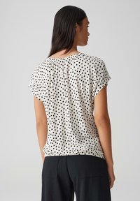 Korte mouw, crèmekleurige blouse met zwarte stippen. Beschikt over een gerimpelde hals en een comfortabele pasvorm, gemaakt van een zachte stof.