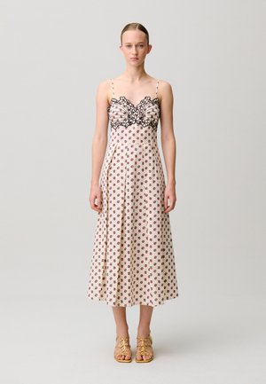 Donna che indossa un vestito midi crema senza maniche con piccoli fiori rossi e dettagli in pizzo nero sul corpetto, abbinato a tacchi gialli con cinturini.