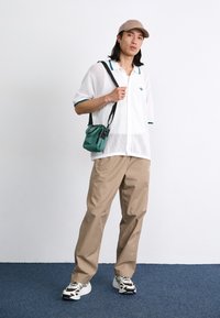 Hvit netting kortermet skjorte med blågrønne detaljer, beige løse bukser og svart-hvite mønstrede joggesko. Liten grønn skulderveske.