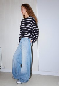 Fiatal nő hosszú hajjal, fekete-fehér csíkos hosszú ujjú ingben, bőszárú világoskék farmerral és ezüst sportcipőben, bent állva.