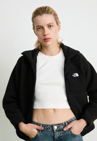Black fleece jacket with a zippered front and high collar, featuring a small side pocket. Worn over a white cropped top and denim jeans.