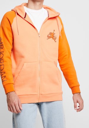 Man wearing peach and orange Nike Air Jordan zip-up hoodie with front pockets and light blue jeans, standing against a light background.