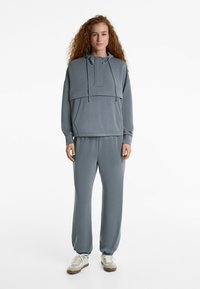 Set de loungewear gri, format din două piese, care include un pulover cu glugă și buzunar frontal, alături de pantaloni joggeri asortați, cu tivuri elastice.