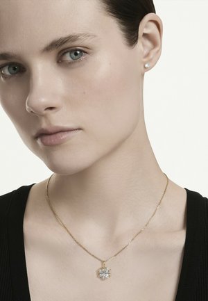 Mujer de piel clara lleva un pequeño pendiente de diamante y un collar de oro con un colgante de diamante en forma de flor, vestida de negro.