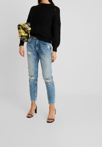 Zwarte gebreide sweater met textuurdetails, gecombineerd met versleten lichtblauwe jeans en zwarte haksandalen. Houdt een geruite gele sjaal vast.