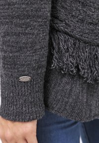 Grau gestrickter Pullover mit einem strukturierten Design, der Fransen am Saum hat und mit einem Metallschild an den Ärmeln versehen ist.