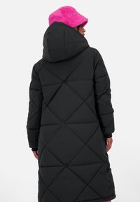 Persona che indossa un lungo cappotto trapuntato nero con cappuccio e un cappello a secchiello morbido di un brillante colore rosa, di spalle contro uno sfondo semplice.