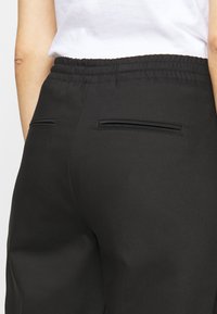 Personne portant un pantalon noir à taille élastique avec deux poches à fentes horizontales à l'arrière et un haut blanc, vue de dos de la taille vers le bas.