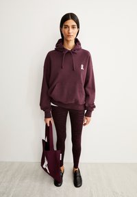 Bordeauxfarbener Hoodie mit einer Vordertasche und weißem Logo, kombiniert mit gemusterten bordeauxfarbenen Leggings. Eine passende Tragetasche wird in einer Hand gehalten.