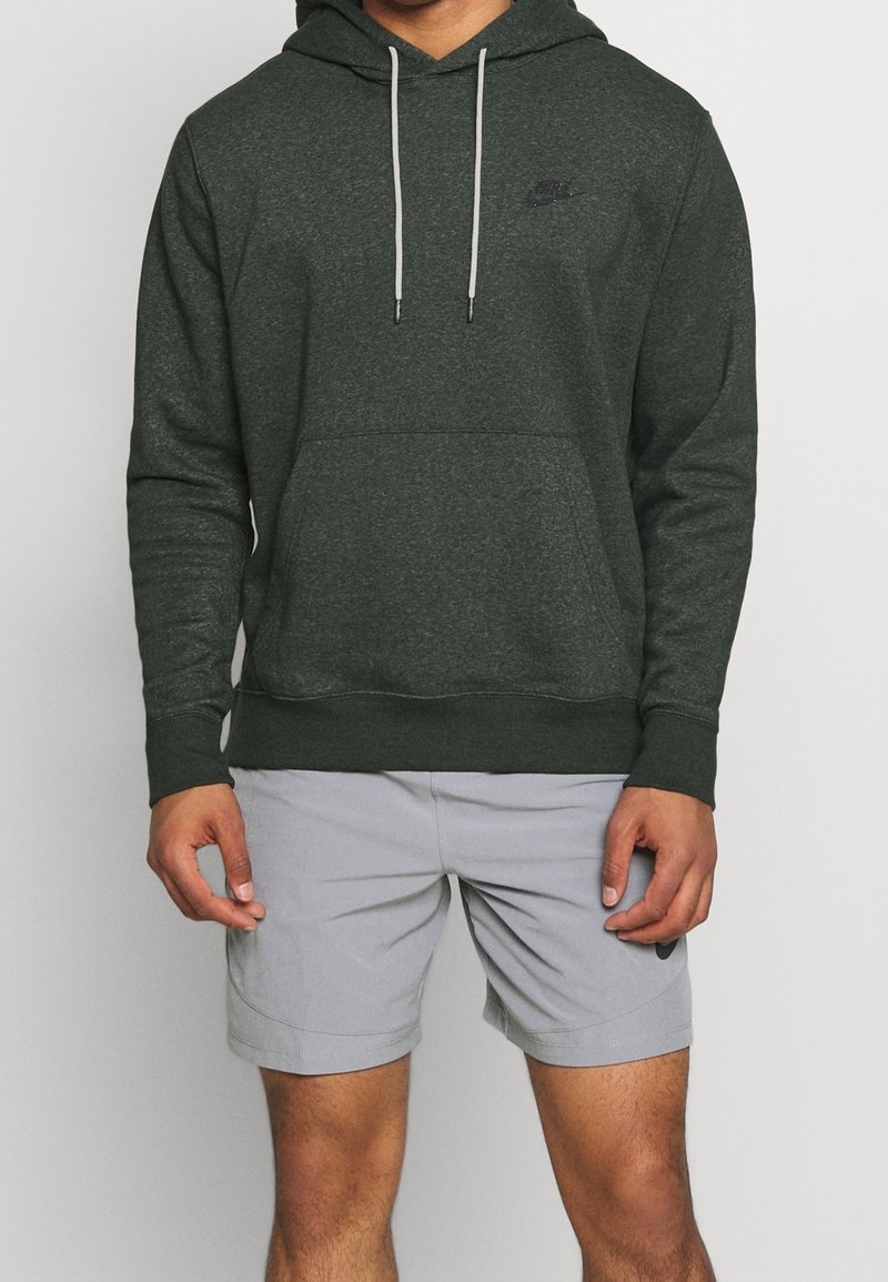 Mörkgrön hoodie med en framficka i kangurustil, grå dragkedjor och en liten svart logotyp på bröstet, i kombination med ljusgrå shorts.