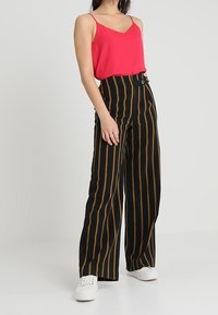 Woman wearing bright pink sleeveless top, high-waisted black pants with brown vertical stripes, and white sneakers, standing against a plain background.