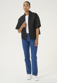 Sort quiltet jakke med brede ærmer, båret over en hvid t-shirt, parret med blå jeans og hvide sneakers. Glat tekstur, minimalistisk design.