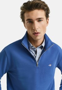 Blaues Zip-Up Sweatshirt mit weicher Textur, Stehkragen, mit kleinem Logo auf der Brust und über einem hellblau gestreiften Hemd getragen.