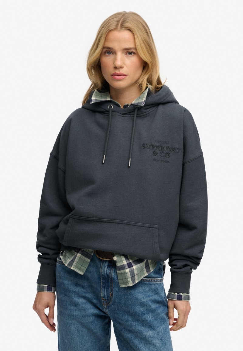 Mujer rubia con sudadera con capucha gris oscuro sobre camisa de cuadros verde y pantalones vaqueros azules, de pie contra un fondo blanco liso.