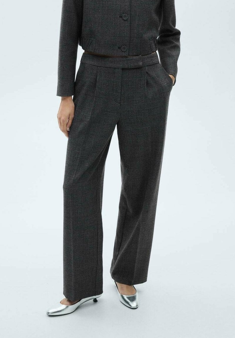 Femme portant un pantalon large à carreaux gris foncé et une veste boutonnée assortie avec des chaussures argentées à talons bas et bouts pointus, mains dans les poches.