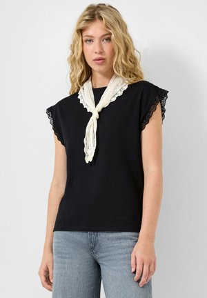 Młoda kobieta o falowanych blond włosach, ubrana w czarny top bez rękawów z koronkowymi brzegami, jasnoniebieskie jeansy i biały szalik związany wokół szyi.