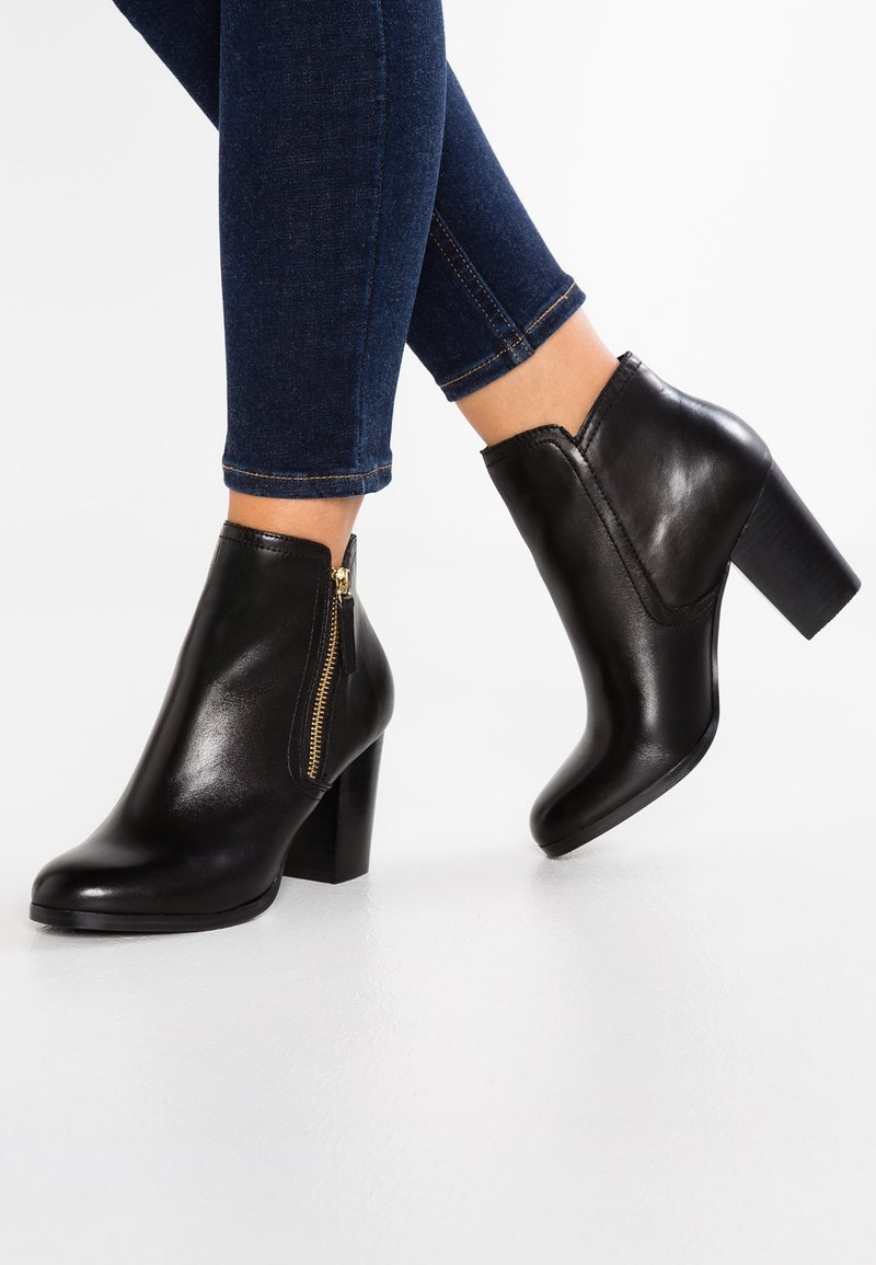 Femme portant un jean skinny bleu foncé et des bottines en cuir noir à talons blocs avec fermetures éclair latérales dorées sur un sol blanc.