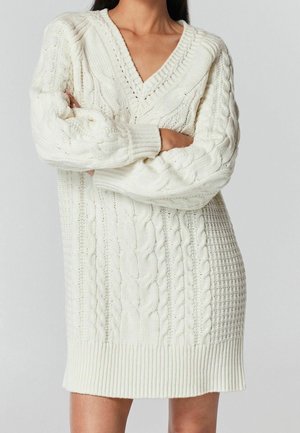 Personne portant une robe-pull blanche en maille torsadée avec col en V, manches longues et motifs texturés, bras croisés sur la poitrine.