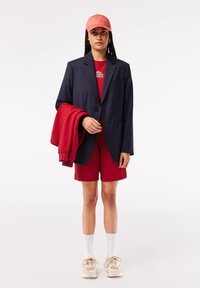 Blazer navy sobre una camiseta de cuello redondo roja, pantalones cortos rojos y una gorra rosa. Zapatillas ligeras con acentos beiges y calcetines blancos. Sosteniendo un sudadera roja.