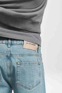 Grauer Sweatshirt, das über einer hellblauen Jeans getragen wird. Die Jeans haben ein Etikett mit "2Y STUDIOS" und einem klassischen Gesäßtaschen-Design.