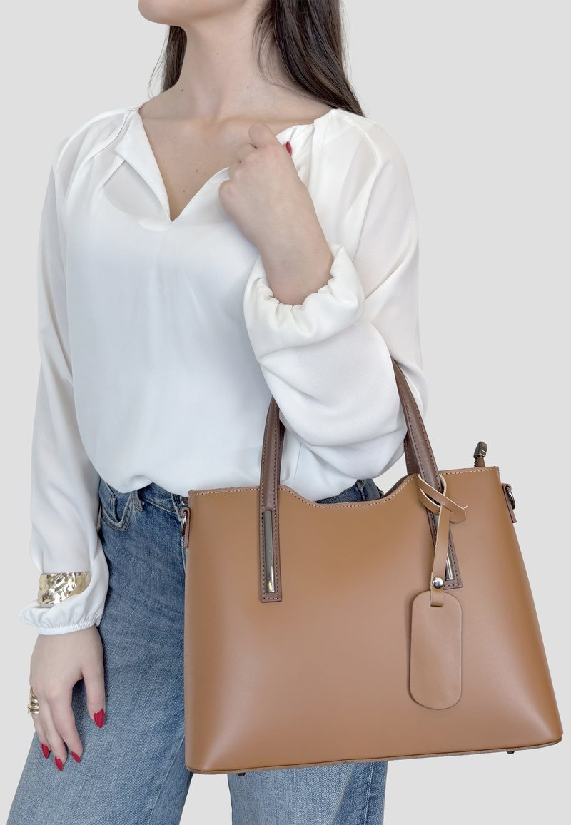 Donna con camicetta bianca e jeans blu che tiene una grande borsa di pelle color cuoio con doppi manici e una targhetta per bagagli.