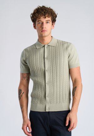 Jeune homme aux cheveux bouclés portant une chemise tricotée à manches courtes vert clair boutonnée, debout devant un fond uni.