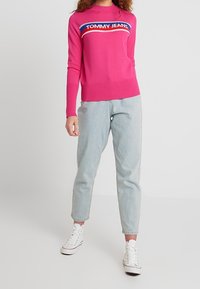 Rosa Strickpullover mit Rundhalsausschnitt und blauer Logoborte. Kombiniert mit hellblauen Jeans und weißen High-Top Sneakers.