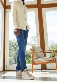 Man in een beige shirt, blauwe spijkerbroek en beige sneakers staat naast een houten stoel met beige kussens bij grote ramen.
