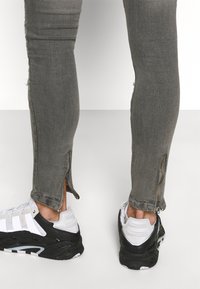 Jeans ajustados grises con desgastes, que cuentan con dobladillos con cremallera, combinados con zapatillas deportivas en blanco y negro. Textura suave y diseño ceñido.