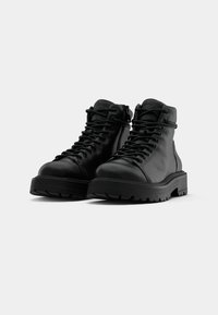 Une paire de bottes en cuir noir à lacets avec des semelles épaisses sur fond blanc, légèrement inclinées vers la gauche.