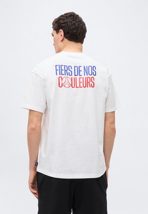 Mężczyzna ubrany w białą koszulkę z niebiesko-czerwonym napisem "FIERS DE NOS COULEURS" i czerwonym okrągłym logo na plecach, czarne spodenki.
