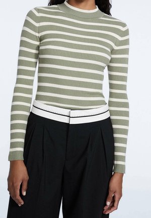 Kvinde iført en tætsiddende olivengrøn og hvid, vandret stribet ribstrikket sweater, stukket ned i højtaljede sorte bukser med et hvidt bælte.