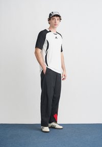 Biała sportowa koszulka z czarnymi akcentami, czarne luźne spodnie z czerwonym trójkątnym detalem oraz białe buty z czarnymi paskami.