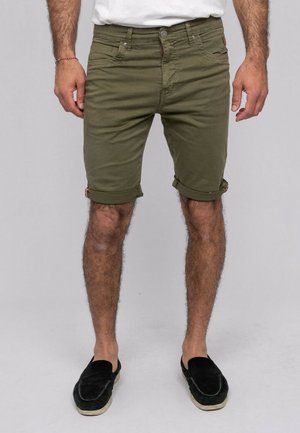 Homme portant un short retroussé vert olive, une chemise blanche, des chaussures noires sans lacets, debout devant un fond uni gris clair.