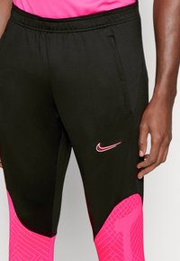 Zwarte sportbroeken met roze accenten, voorzien van een gestructureerd patroon op het onderbeen, elastische tailleband en een rood Nike-logo.