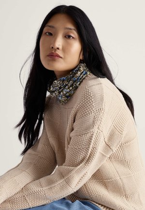 Bež pulover iz pletenine s teksturiranim vzorcem, kombiniran z zelenim cvetličnim rutkom za vrat z modrimi poudarki. Mehki materiali in sproščen kroj.