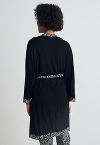 Čierny plášť s dlhými rukávmi, vzorovanými manžetami a zodpovedajúcim opaskom. Má jemnú štruktúru a uvoľnený strih, nosený cez vzorované nohavice.