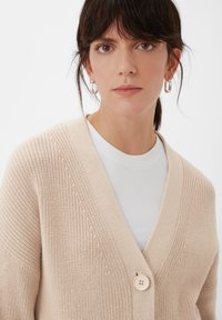 Beige gestrickte Strickjacke mit V-Ausschnitt, die mit einem einzigen Knopf verschlossen wird und ein strukturiertes Muster aufweist. Über einem weißen gerippten Oberteil getragen.