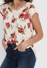 Top a maniche corte di colore crema con motivo floreale rosso e dettagli verdi, con scollatura rotonda e spacchi laterali, abbinato a jeans blu scuro.