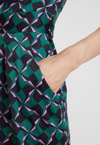Mekko, jossa on geometrinen kuvio vihreässä, mustassa ja vaaleanpunaisessa, ja sivulla näkyy tasku. Kankaan pinta on sileä.